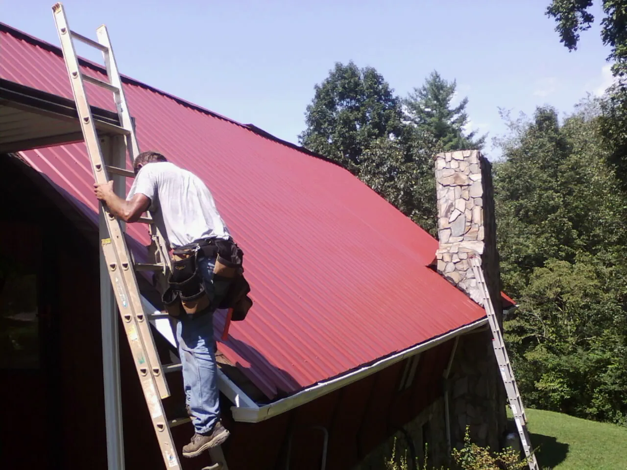 Roofer on a ladder beside a red metal roof and stone chimney — Soffit & Fascia Repair by Roofing Brothers in Eagle